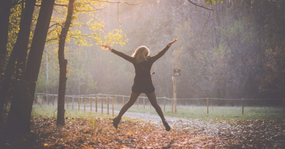 Person leaps with arms outstretched on a leaf-covered forest path, bathed in golden autumn sunlight.