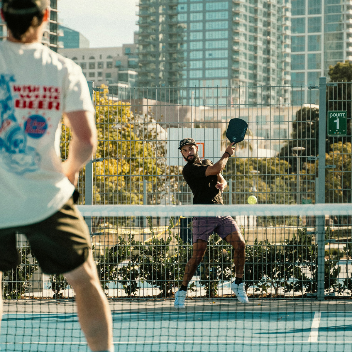 Two men playing pickleball on an outdoor court, with tall buildings and trees in the background. 