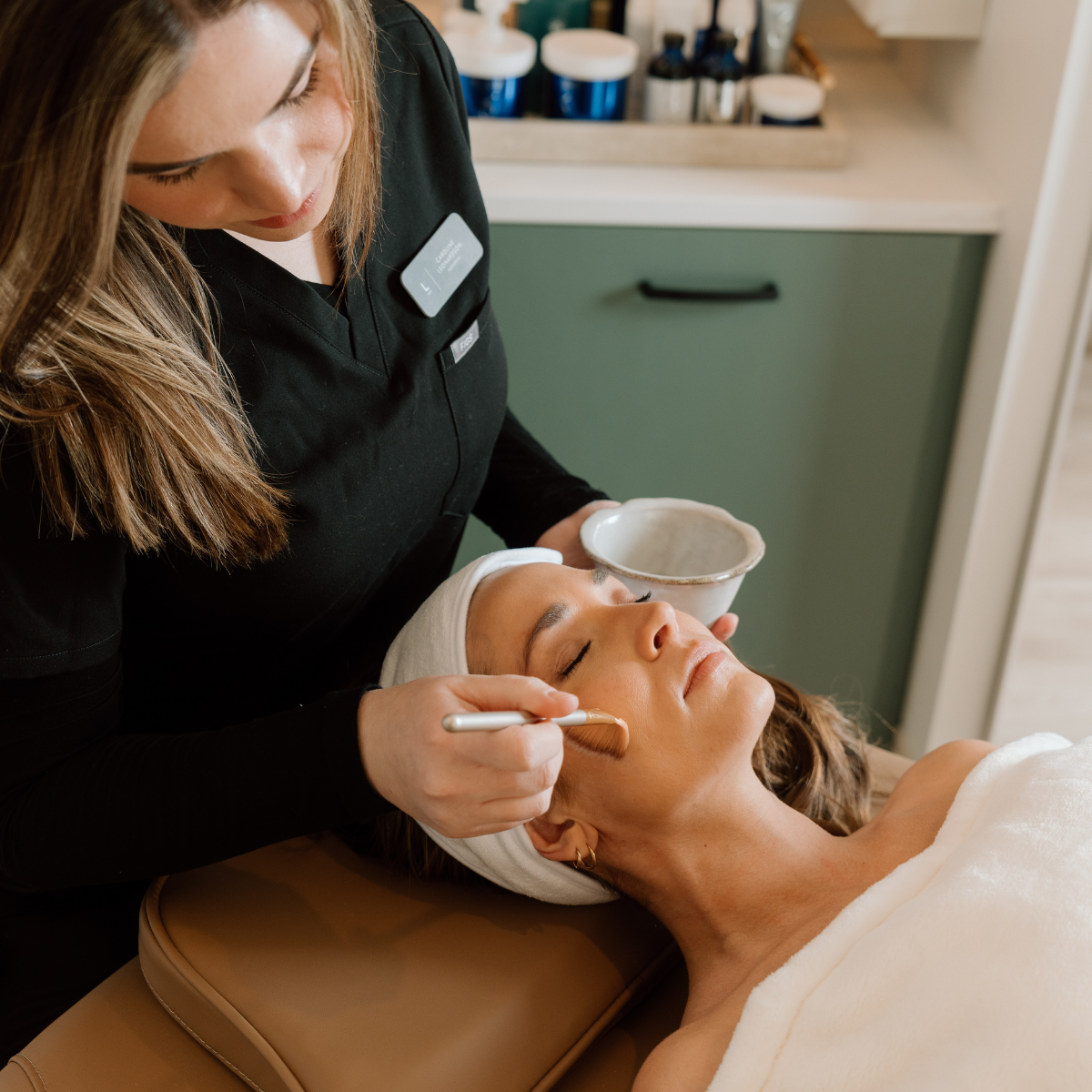 A serene spa scene with a beautician applying a facial mask to a relaxed client's face.