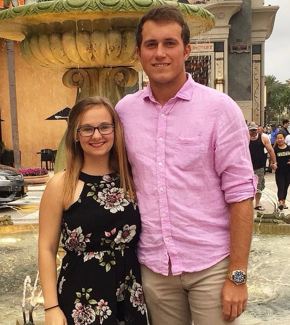 A petite woman wearing a flower-print summer dress stands arm-in-arm with a tall man in a pink shirt and tan pants in front of a fountain. 
