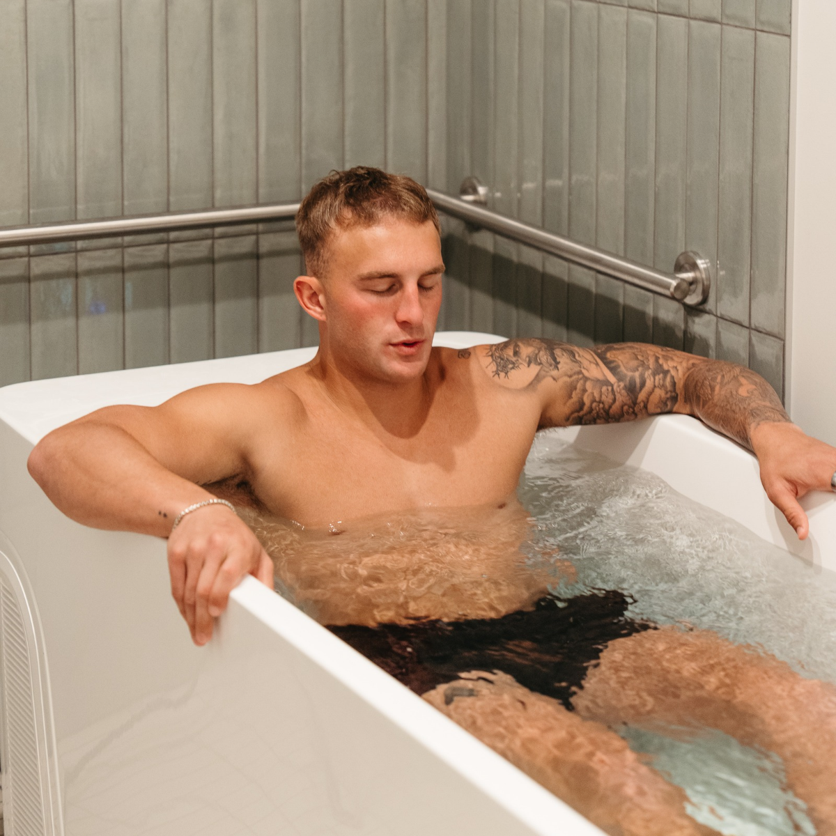 A man relaxes with eyes closed while lying in a white bathtub filled with cold water in a calm and restorative contrast therapy room. 