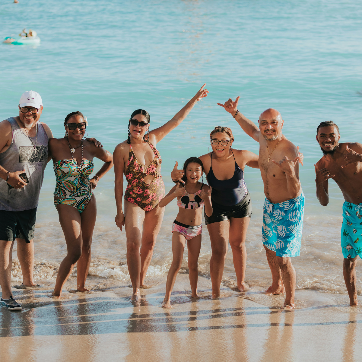 Group of seven people of various ages smiling and posing at the beach with ocean waves behind them.