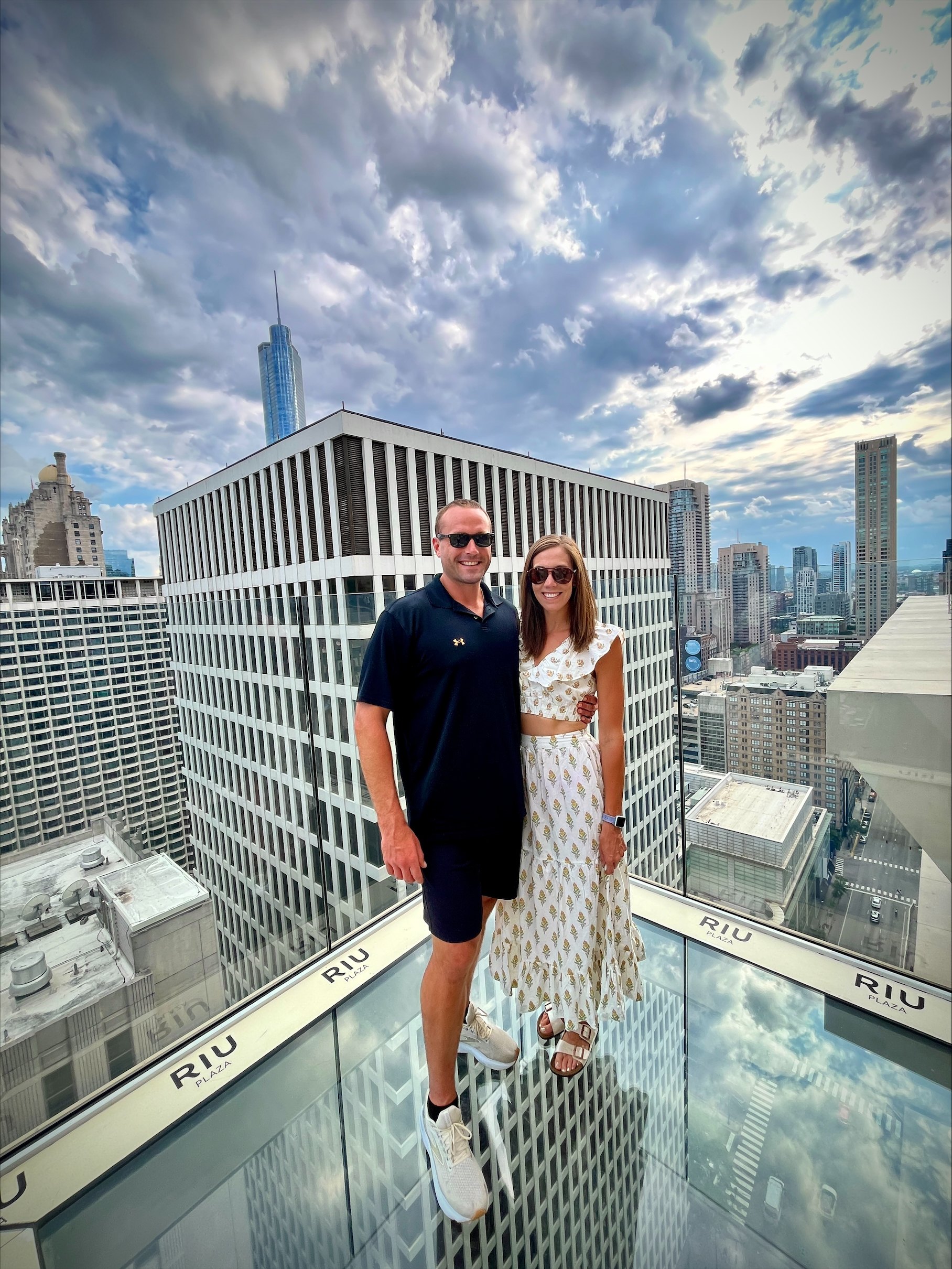 A man wearing a black polo shirt and black shorts stands on top of the Skydeck in Chicago with a woman in a two - piece brown and white flower dress.