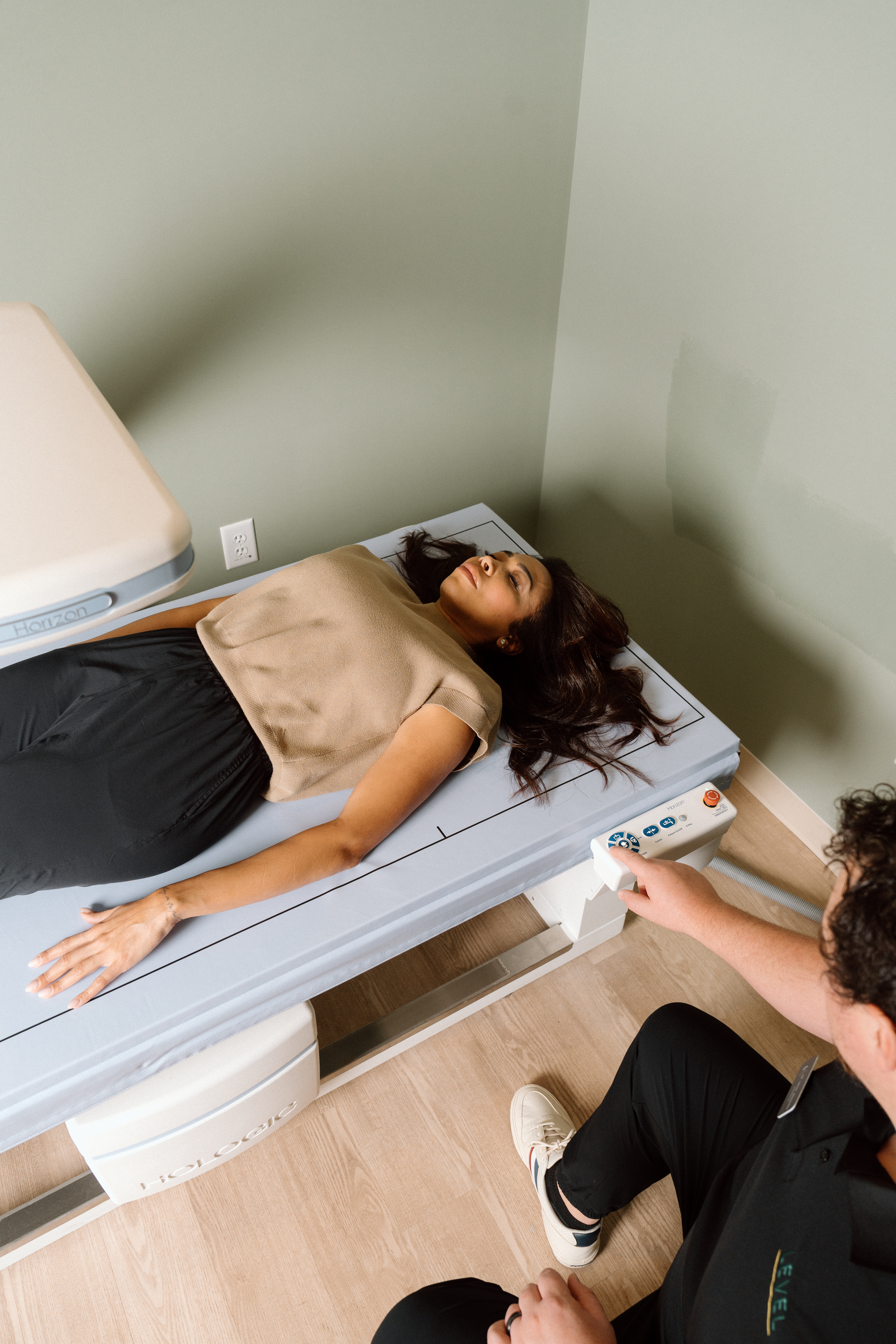 A patient dressed in black pants and a beige blouse lays on their back on a DXA scan table while a technician performs the assessment.