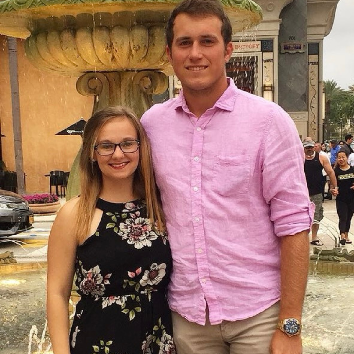 A petite woman wearing a flower-print summer dress stands arm-in-arm with a tall man in a pink shirt and tan pants in front of a fountain. 