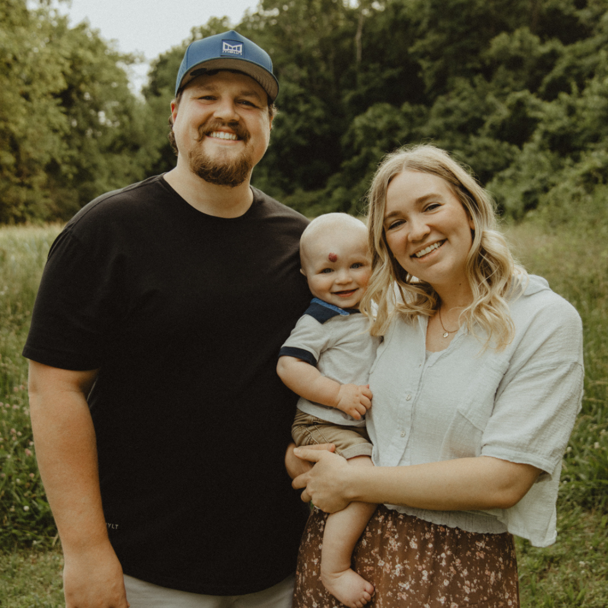 A couple smiles while holding a baby and standing in a field with lush green trees in the background. 