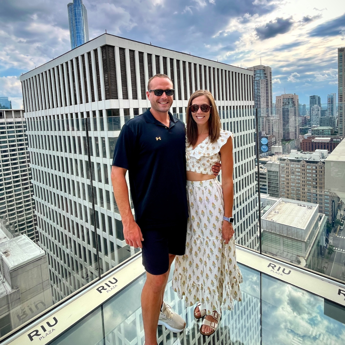 A man wearing a black polo shirt and black shorts stands on top of the Skydeck in Chicago with a woman in a two - piece brown and white flower dress.