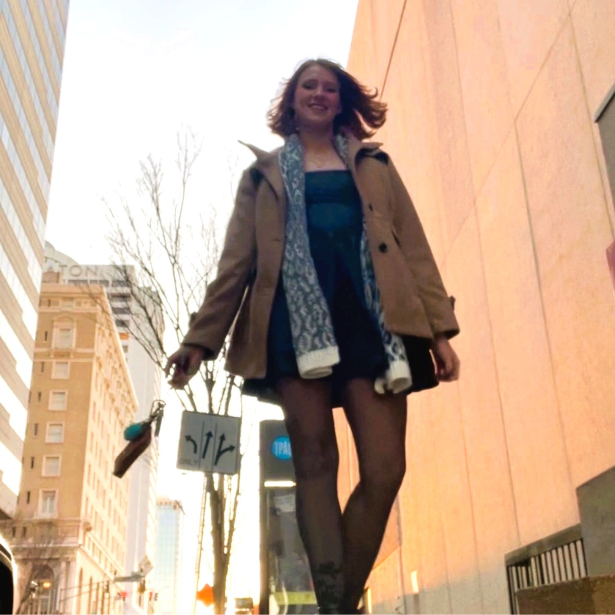 Photo from ground level of woman walking along sidewalk, wearing a hip-length tan pea coat, blue and white scarf, and dark miniskirt.
