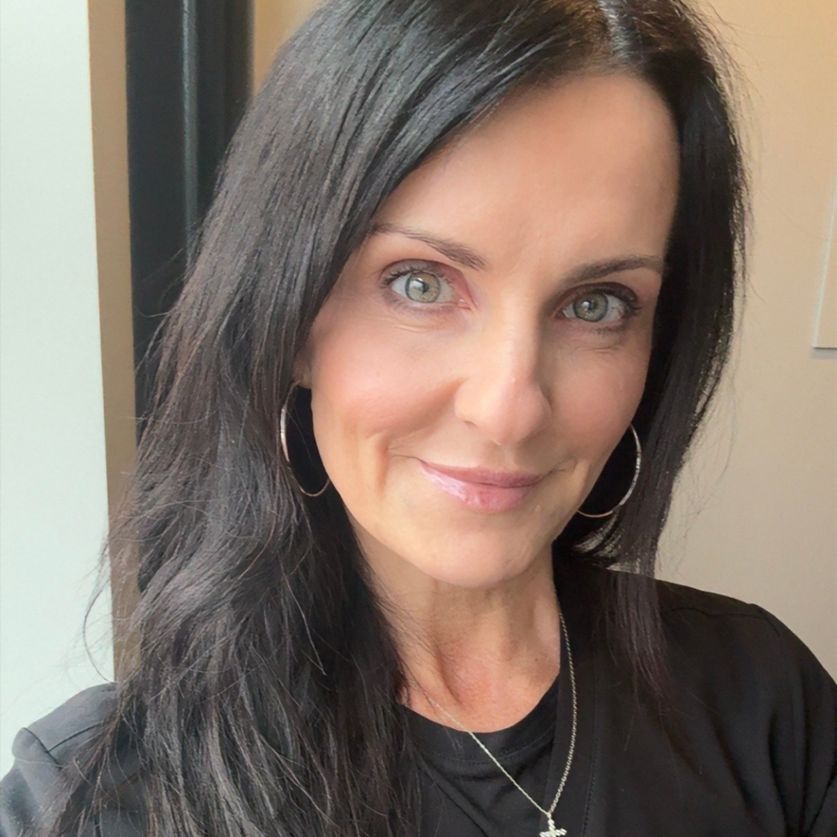 A woman with long dark hair and green eyes, wearing a black top, smiles for a selfie. 