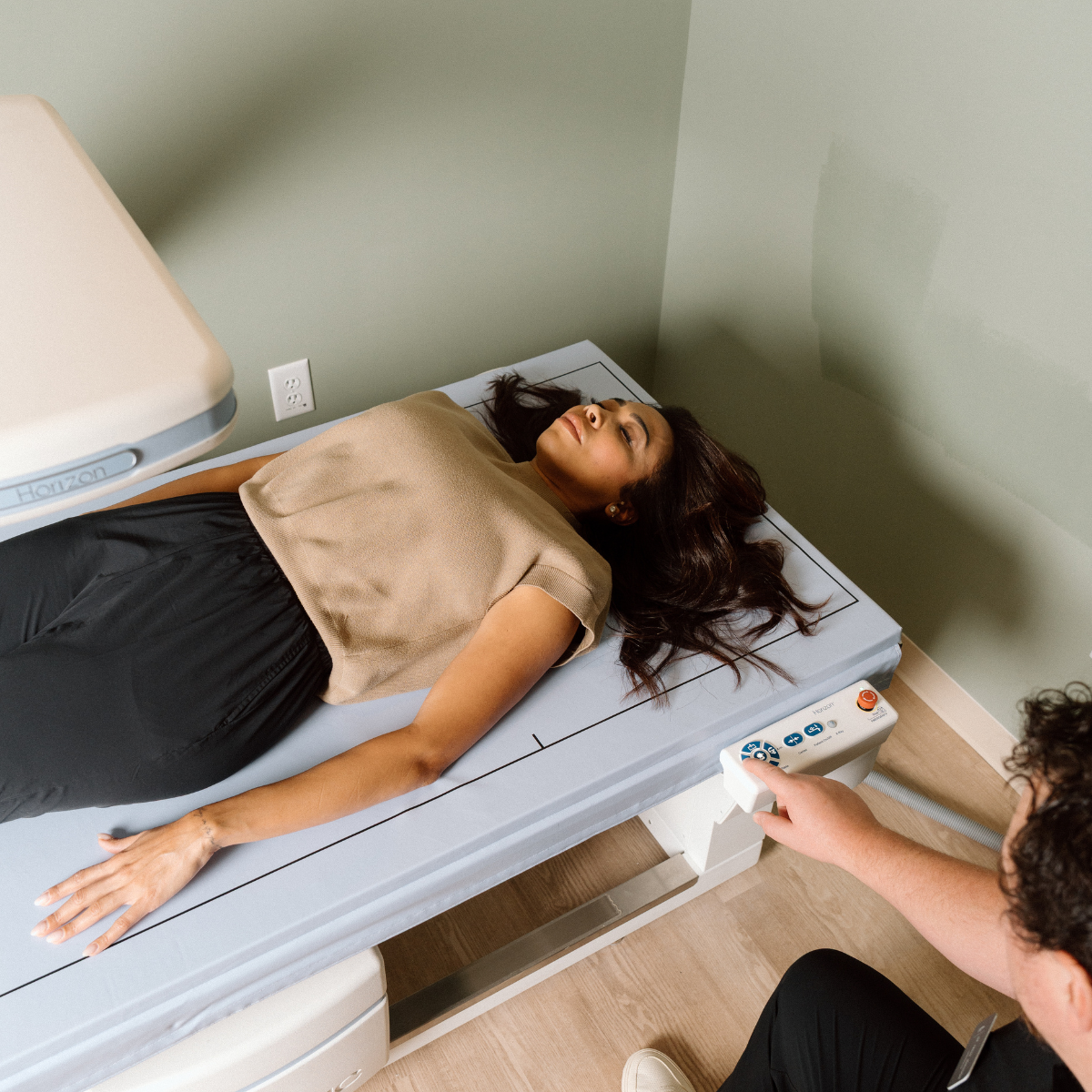 A patient dressed in black pants and a beige blouse lays on their back on a DXA scan table while a technician performs the assessment.