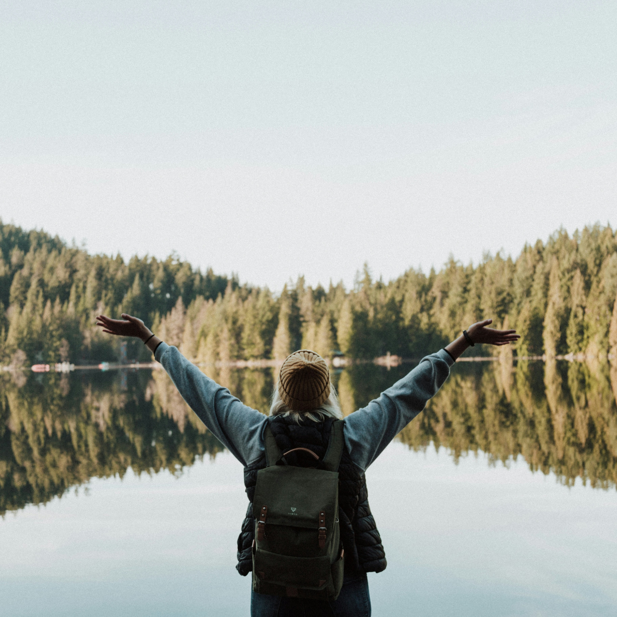Woman with her arms stretched wide, facing a calm body of water surrounded by trees.