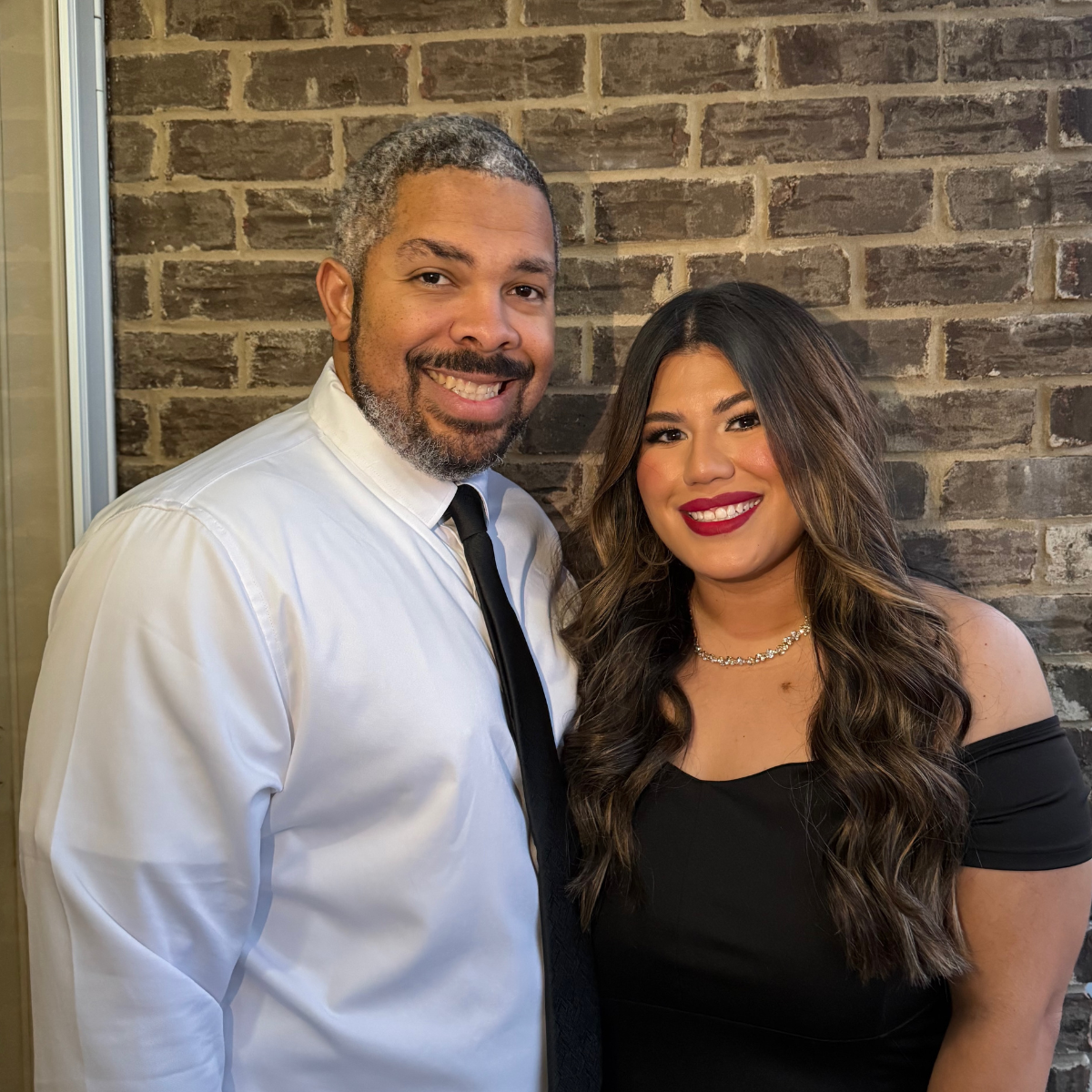 Smiling couple dressed in cocktail attire stand in front of an exterior brick wall with their arms around each other’s waists.