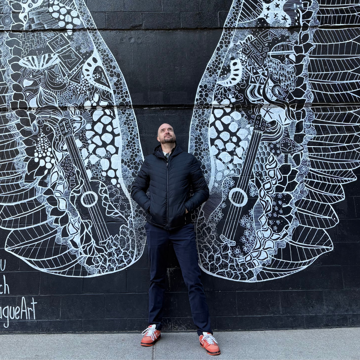 A man stands beneath a giant black-and-white angel-wing mural on a dark brick wall, surrounded by intricate patterns and street-art text.