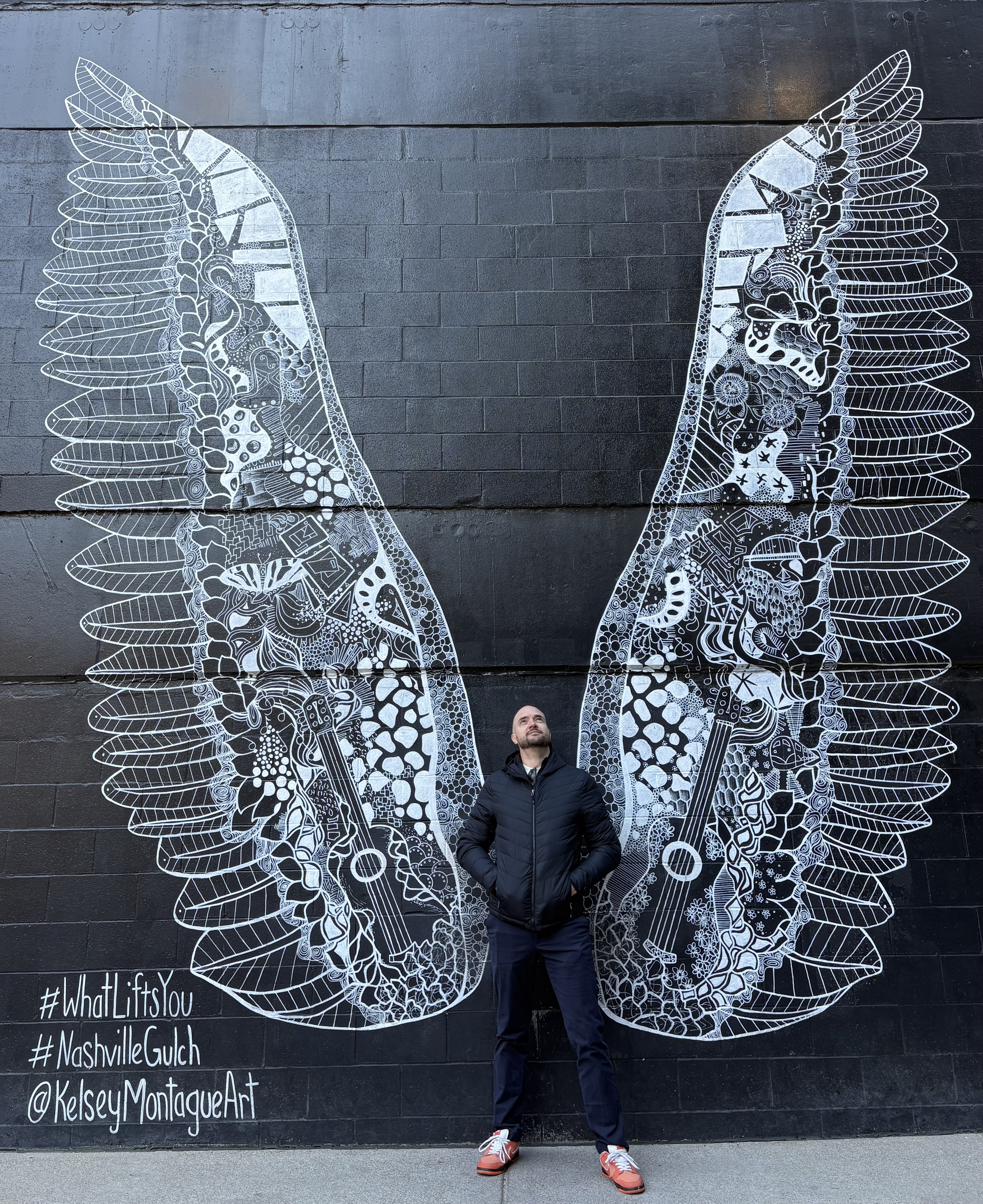 A man stands beneath a giant black-and-white angel-wing mural on a dark brick wall, surrounded by intricate patterns and street-art text.