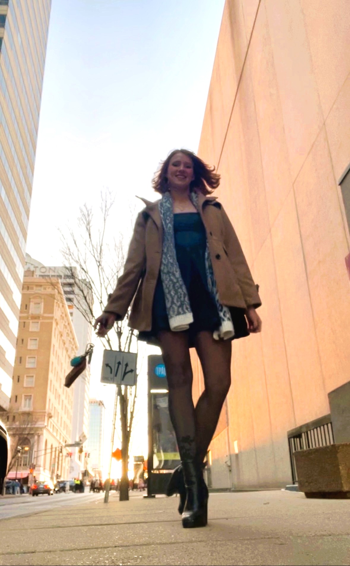 Photo from ground level of woman walking along sidewalk, wearing a hip-length tan pea coat, blue and white scarf, and dark miniskirt.