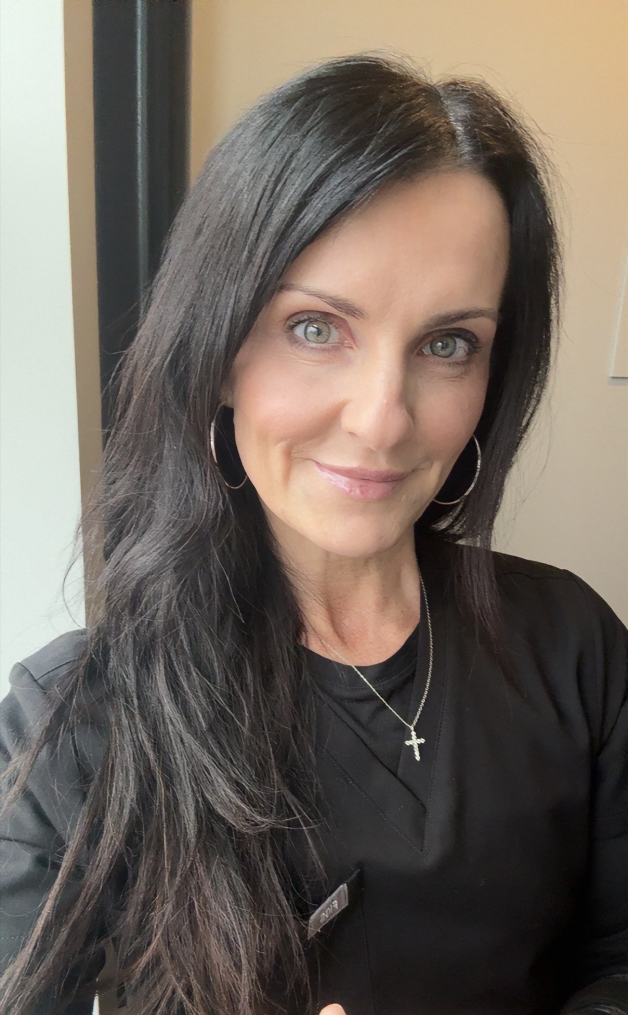 A woman with long dark hair and green eyes, wearing a black top, smiles for a selfie. 