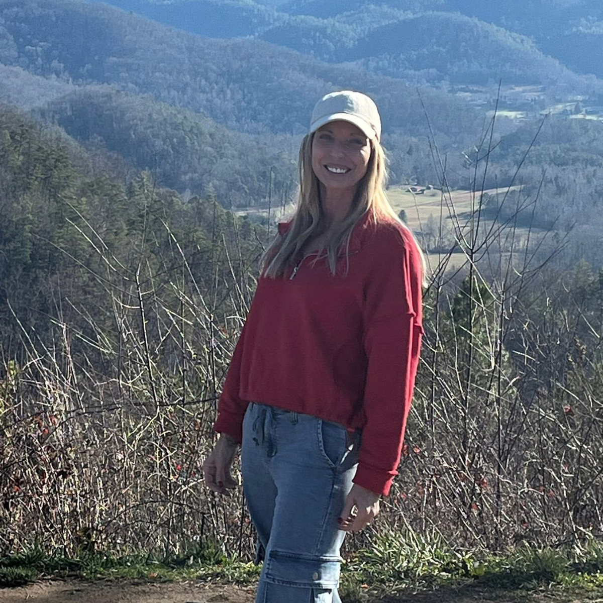 A woman with long blond hair, wearing a red sweater, jeans, and white hat, stands smiling in front of a scenic, mountainous landscape.
