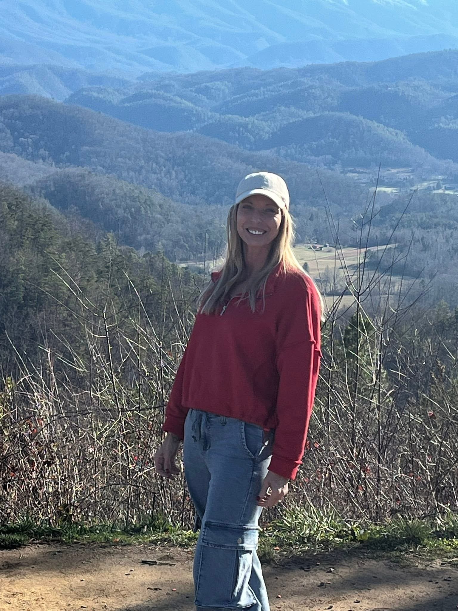 A woman with long blond hair, wearing a red sweater, jeans, and white hat, stands smiling in front of a scenic, mountainous landscape.