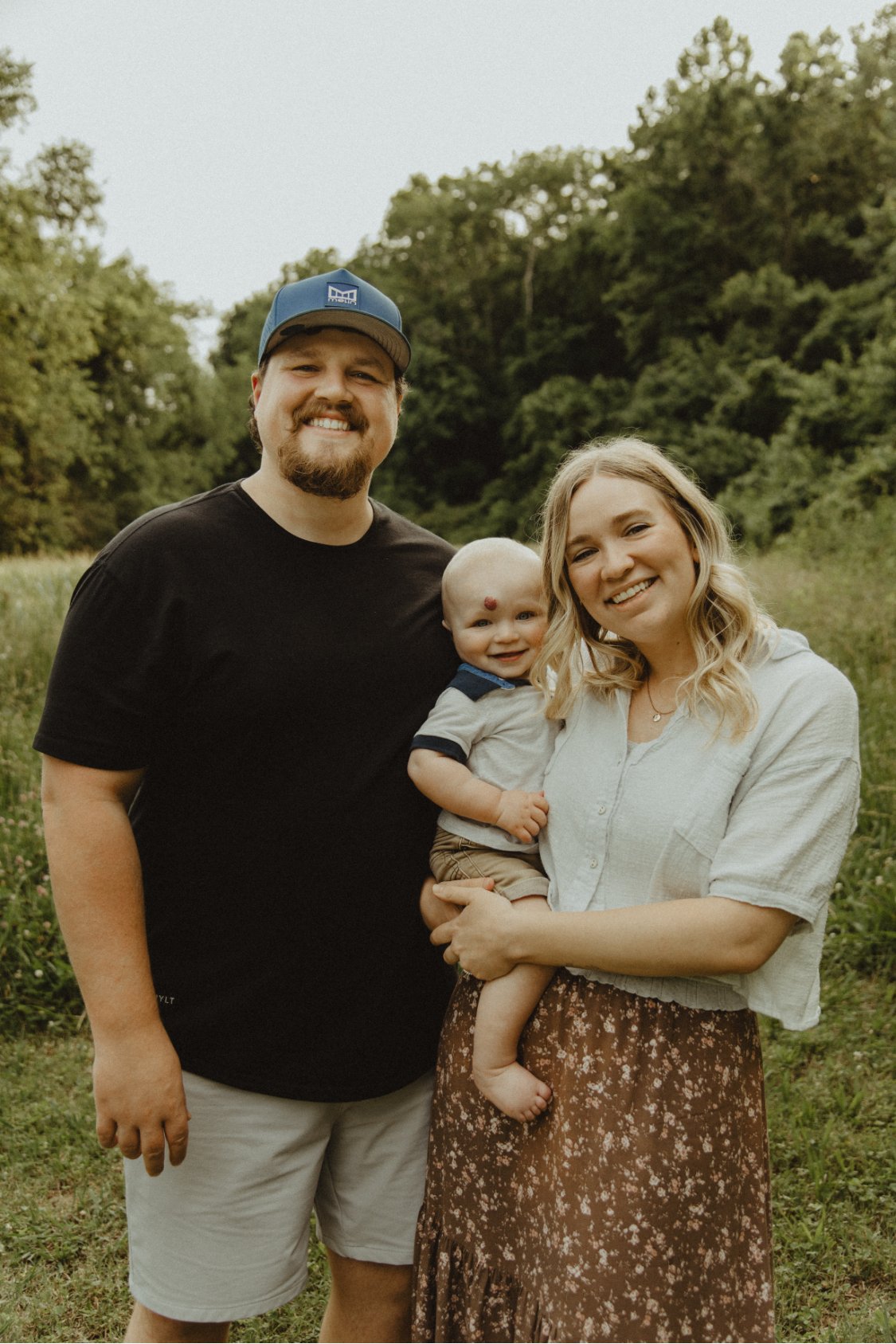 A couple smiles while holding a baby and standing in a field with lush green trees in the background. 