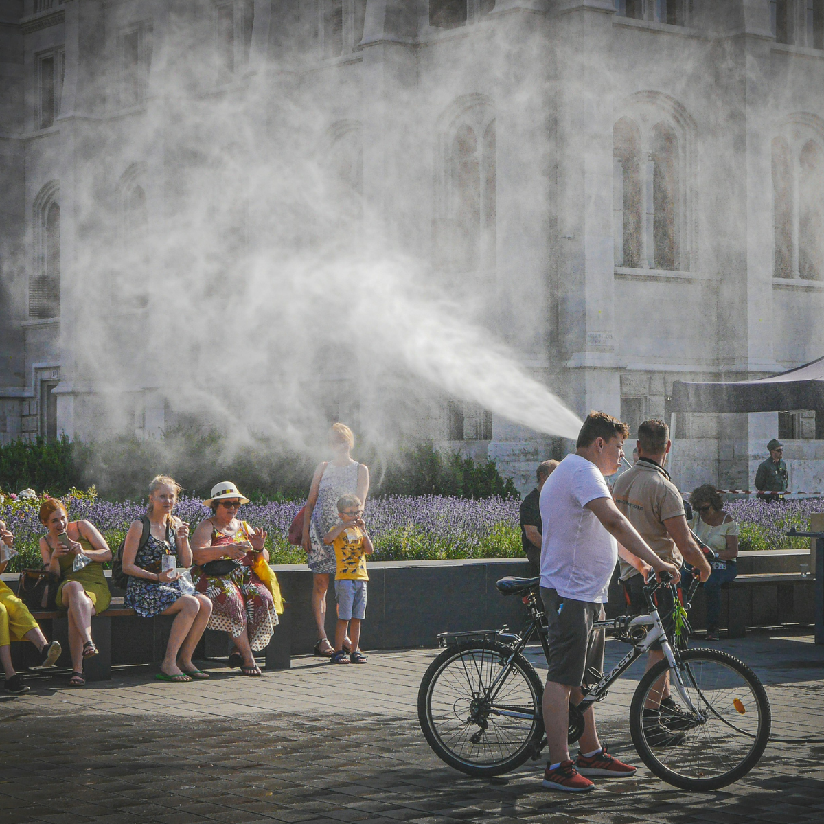 Street scene of people sitting on benches on a hot summer day with a cyclist passing through a cooling mist. 