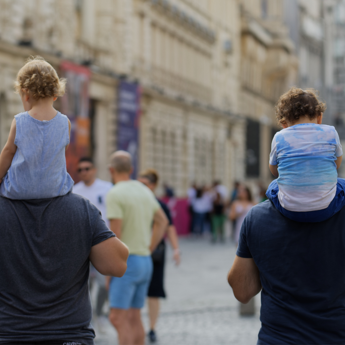 Two men carry children on their shoulders while walking down a bustling city street. Historic buildings line the background in a lively and joyful scene. 