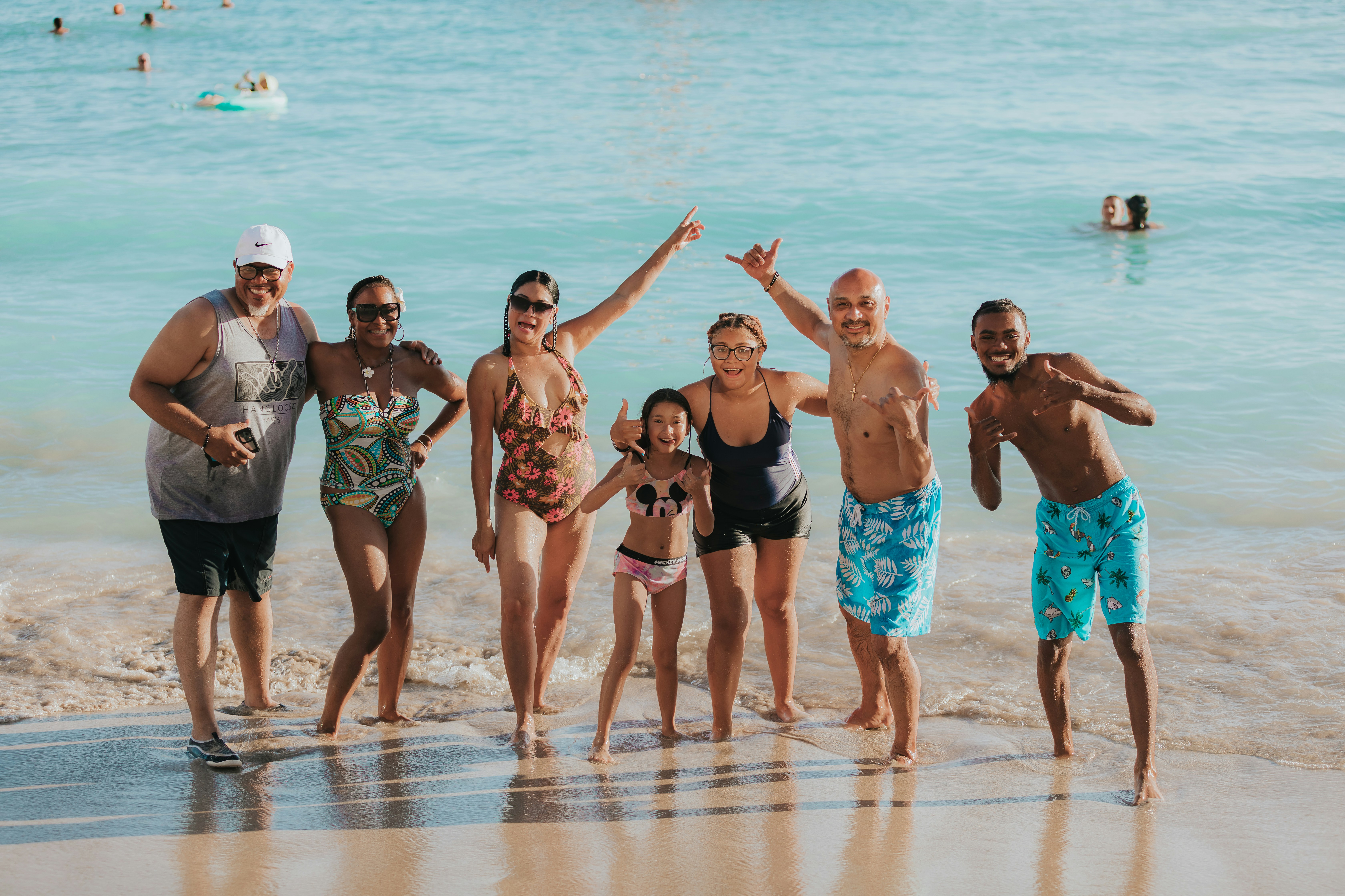 Group of seven people of various ages smiling and posing at the beach with ocean waves behind them.