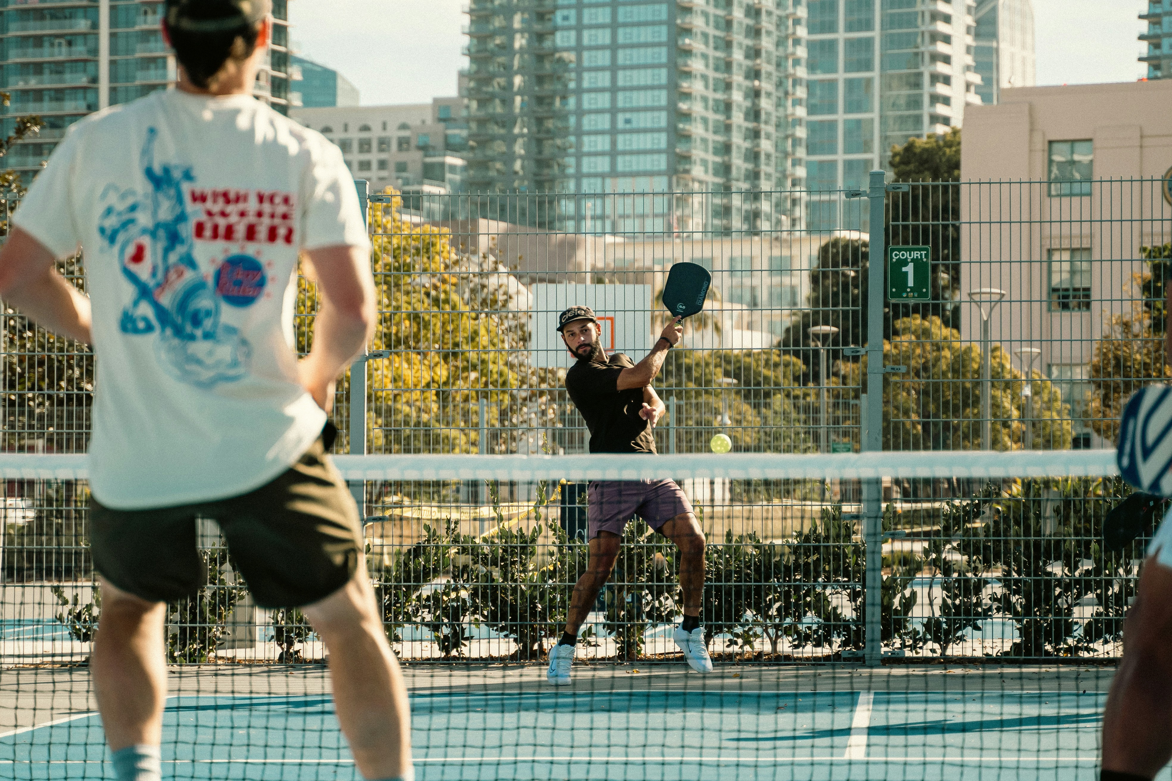 Two men playing pickleball on an outdoor court, with tall buildings and trees in the background. 