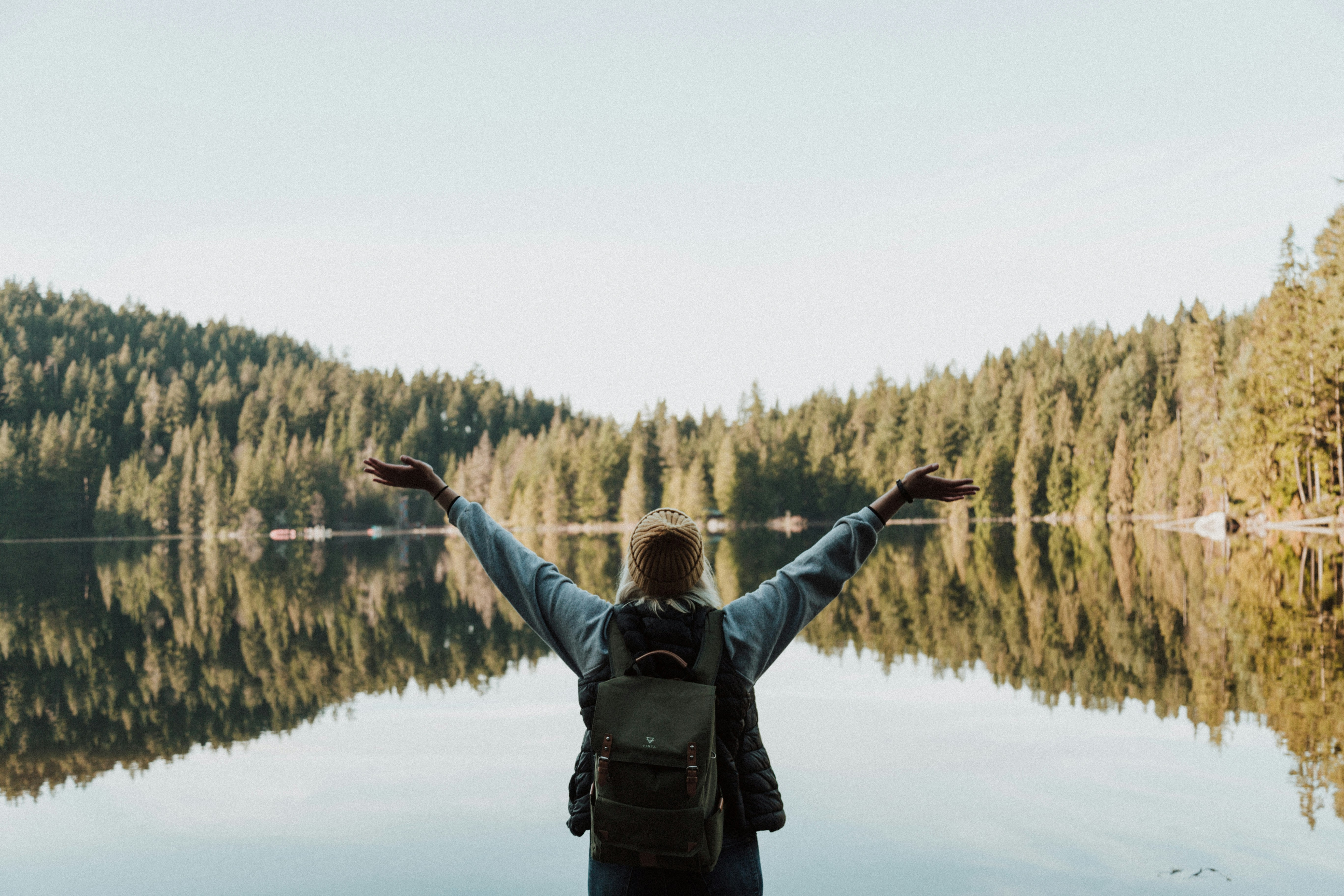 Woman with her arms stretched wide, facing a calm body of water surrounded by trees.