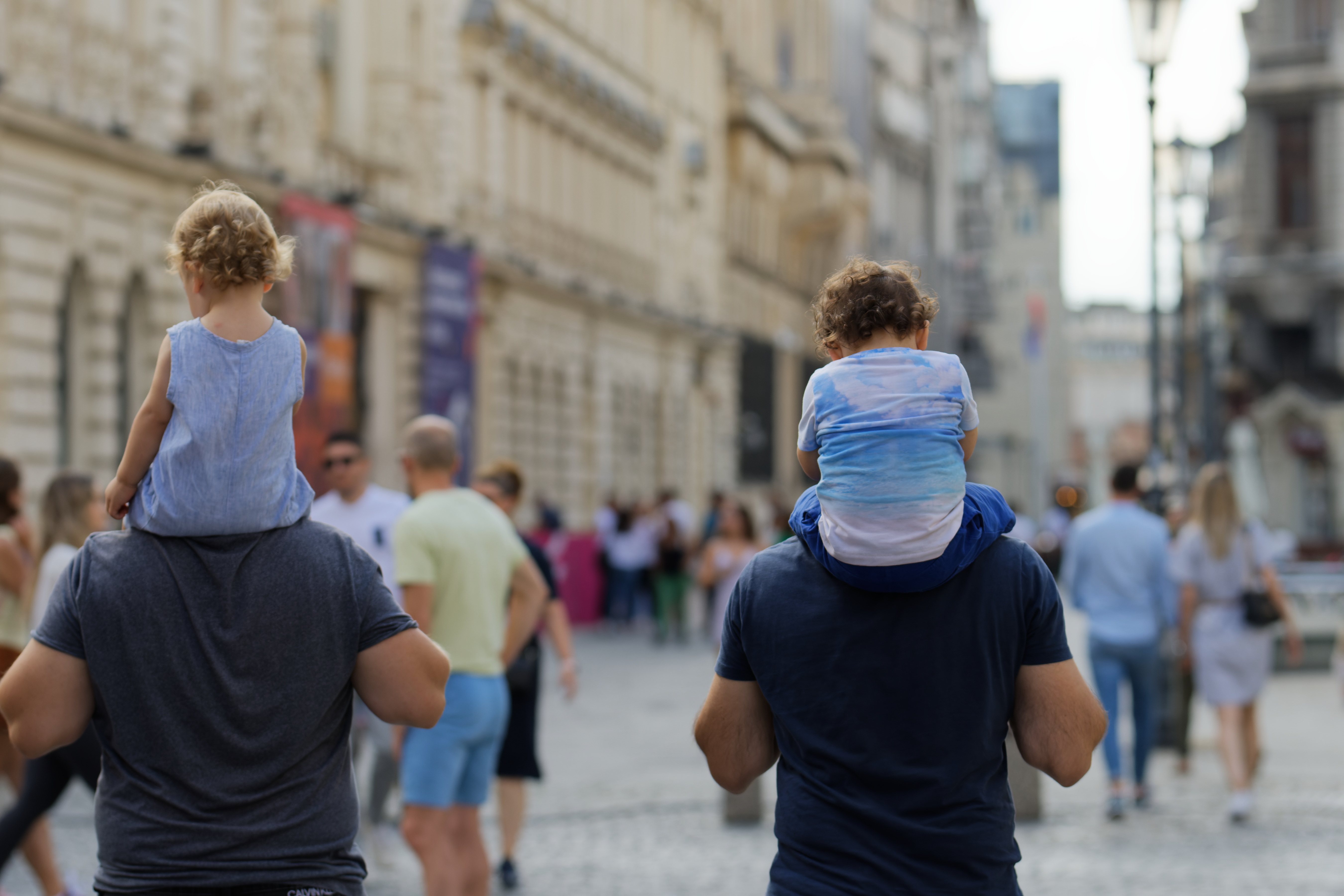 Two men carry children on their shoulders while walking down a bustling city street. Historic buildings line the background in a lively and joyful scene. 